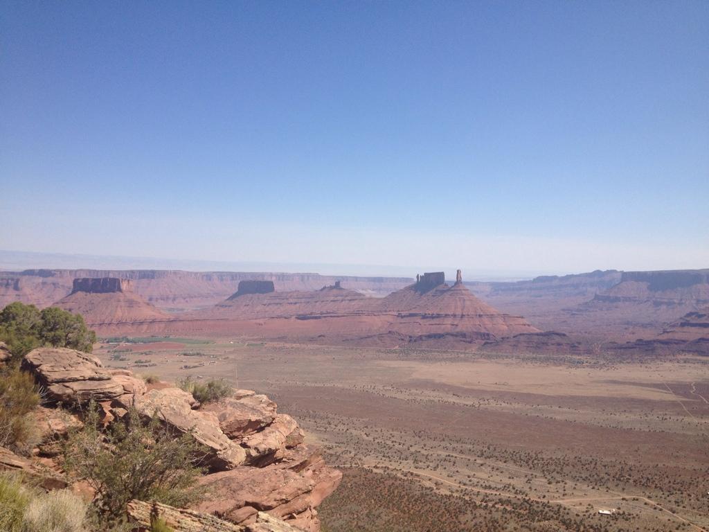 A panoramic view of a desert landscape featuring reddish rock formations and mesas under a clear blue sky. The foreground includes shrubs and rocky outcrops, while distant mesas stretch across the horizon, showing varying heights and formations. Porcupine Rim mountain bike trail.