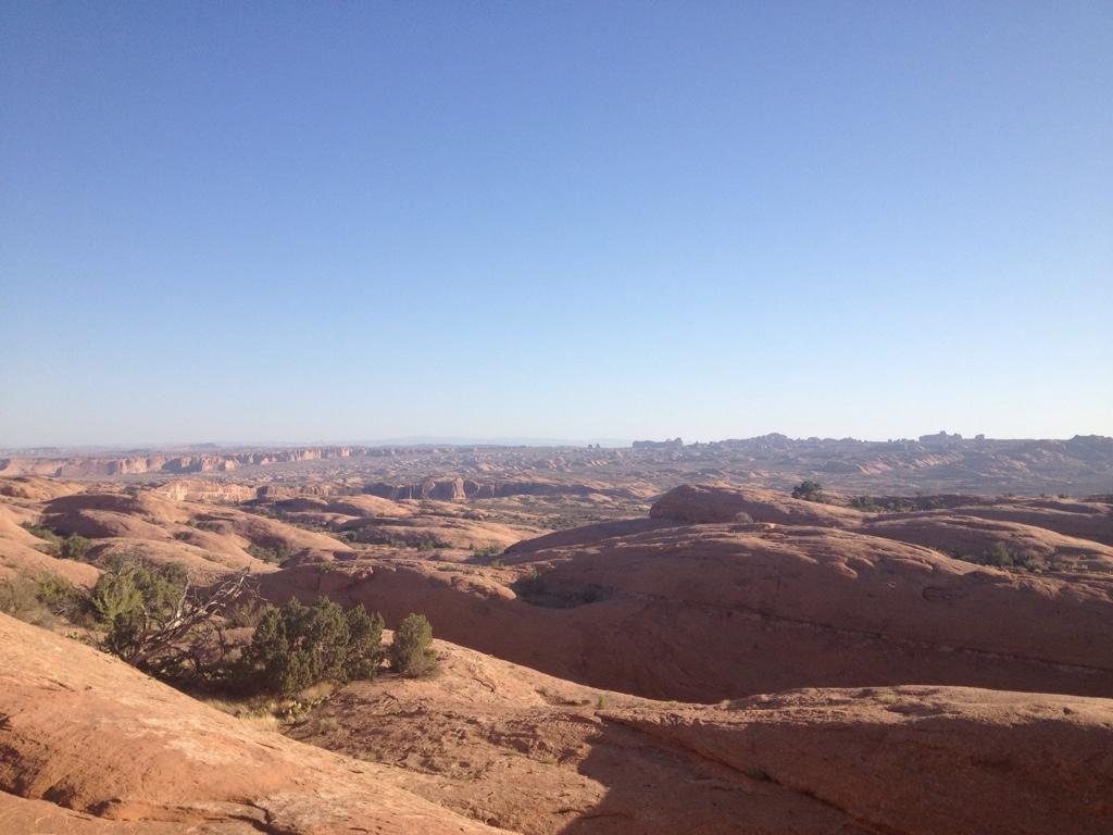 A panoramic view of a desert landscape featuring red rock formations and hills under a clear blue sky, with sparse vegetation in the foreground. Slickrock mountain bike trail.
