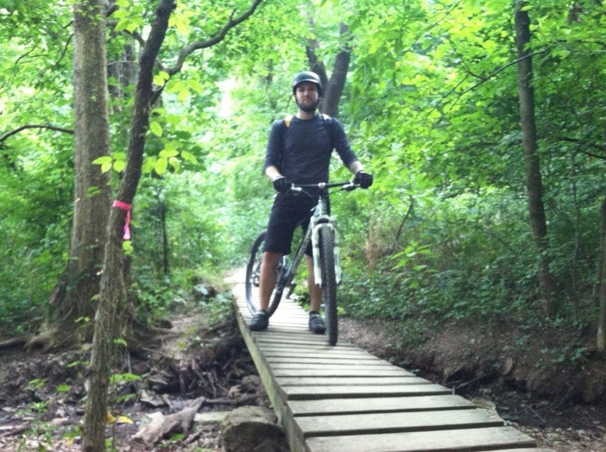 A person wearing a helmet and gloves stands beside a mountain bike on a wooden bridge in a lush green forest. The surrounding area is densely wooded, with trees and foliage creating a vibrant, natural setting. Frick Park mountain bike trail.