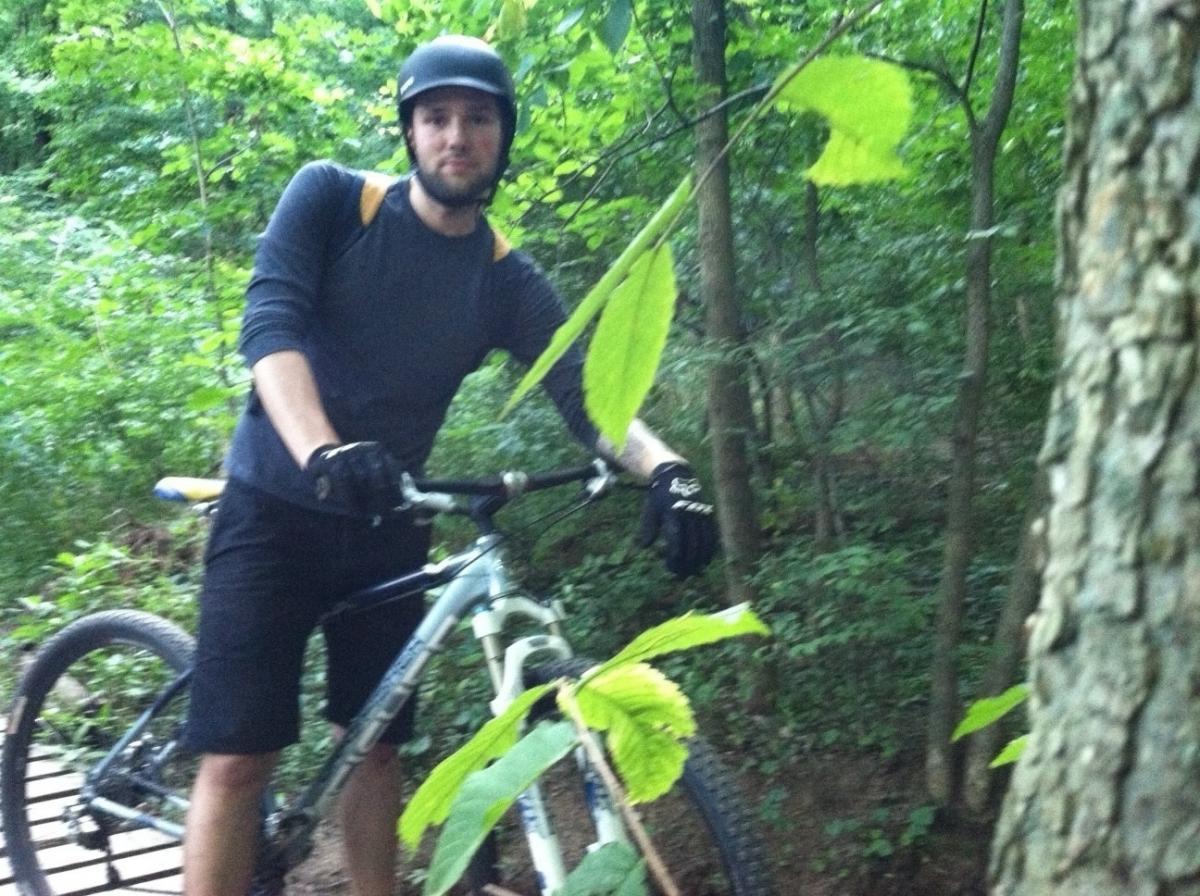 A mountain biker wearing a helmet and gloves stands next to his bike on a forest trail, surrounded by green foliage and trees. Frick Park mountain bike trail.