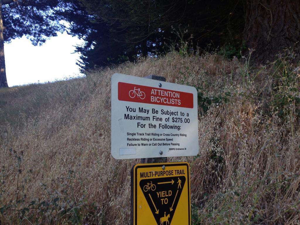A warning sign for bicyclists indicating potential fines of up to $275 for specific offenses, including reckless riding and failure to alert others when passing. The sign also emphasizes adherence to trail rules. Redwood Regional Park mountain bike trail.