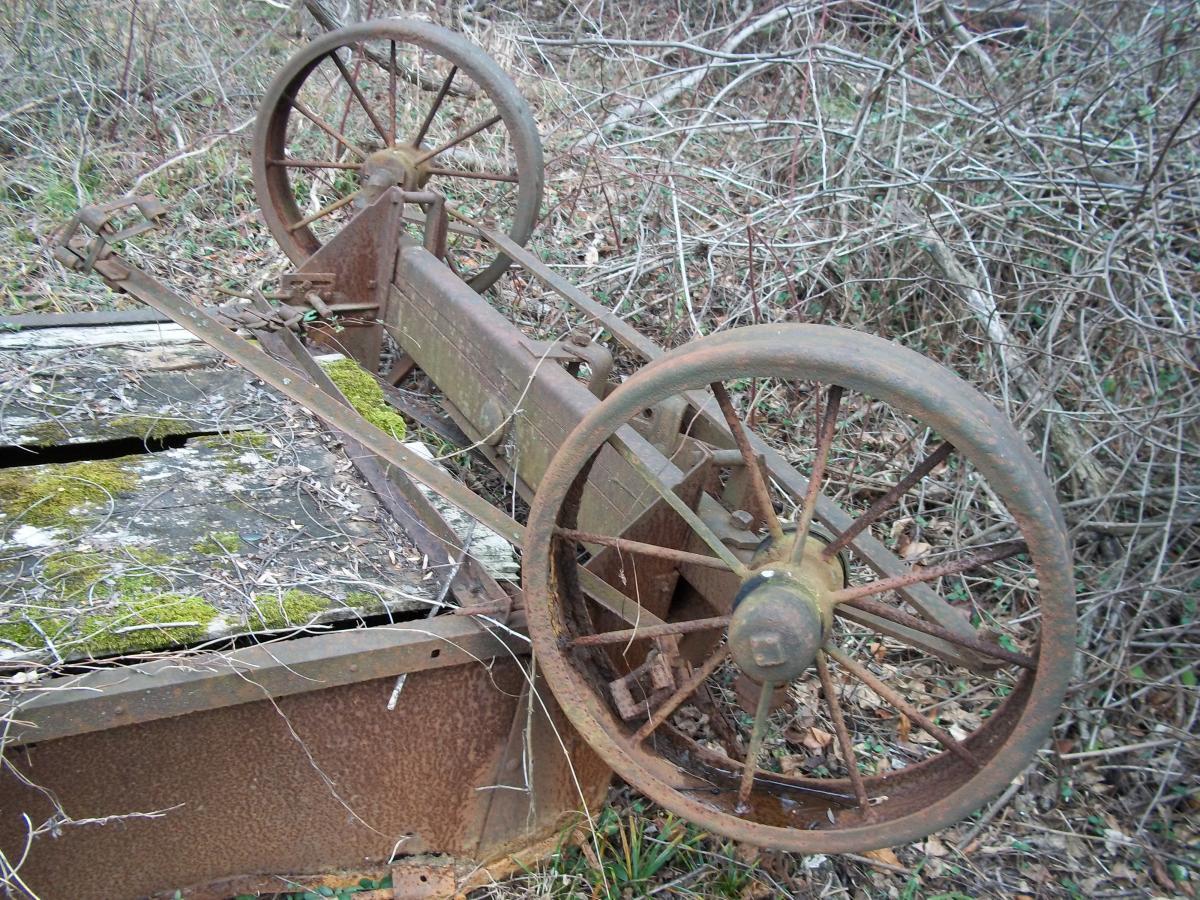 An old, rusty farm implement with two large, weathered wheels, partially covered in vegetation. The implement appears to be overturned, revealing a metal frame and remnants of mechanical attachments, set against a backdrop of overgrown grass and shrubs. Patapsco Valley State Park (Avalon Area) mountain bike trail.
