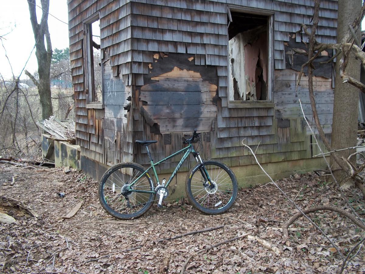 A green mountain bike leaning against the exterior of an abandoned wooden house with weathered shingles. The building shows signs of decay, including broken windows and peeling paint, with scattered leaves and debris on the ground surrounding the structure. Trees and underbrush are visible in the background. Patapsco Valley State Park (Avalon Area) mountain bike trail.