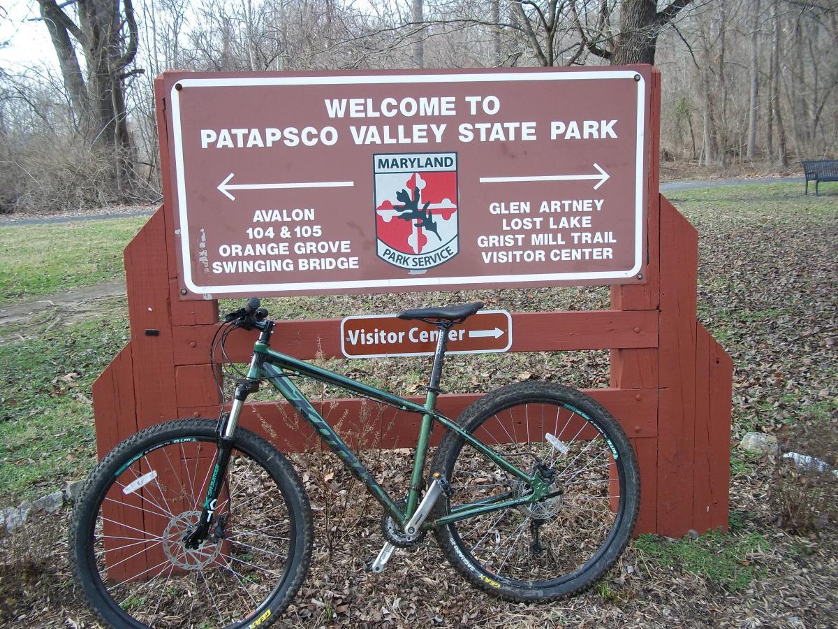 Alt text: A green mountain bike is parked next to a wooden sign that reads "Welcome to Patapsco Valley State Park." The sign lists nearby locations such as Avalon, Glen Artney, and Lost Lake, along with a directional arrow and a visitor center. The background includes grass and trees, suggesting an outdoor environment. Patapsco Valley State Park (Avalon Area) mountain bike trail.