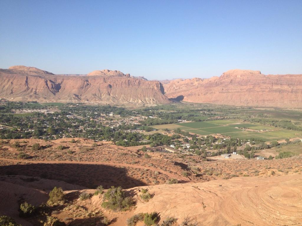 A panoramic view of a valley surrounded by rocky hills, featuring a small town nestled among green fields and trees. The landscape showcases varying shades of brown and green under a clear blue sky. Slickrock mountain bike trail.