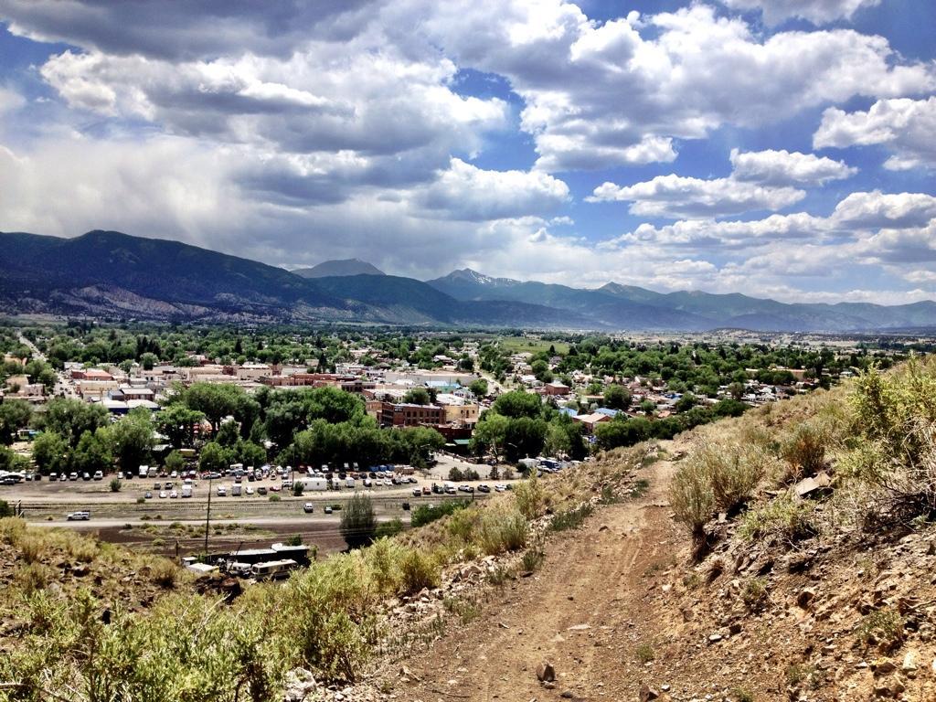 A scenic view from a hillside overlooking a small town surrounded by greenery, with mountains in the background and a partly cloudy sky. The image captures a dirt path leading down the hill, showcasing residential buildings and parked cars in the foreground. Frontside mountain bike trail.