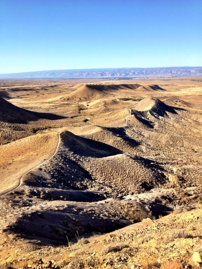 A panoramic view of a barren landscape featuring rolling hills and rugged terrain under a clear blue sky. The ground is primarily dry and rocky, with subtle shadows and textures visible on the hills. In the distance, a mountain range can be seen, adding depth to the horizon. Zippety Do Dah mountain bike trail.