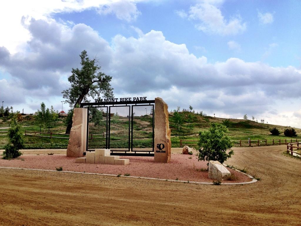 Alt text: Entrance gate of Valmont Bike Park, featuring large stone pillars and a metal sign, set against a backdrop of grassy hills and a cloudy sky. Valmont Bike Park mountain bike trail.