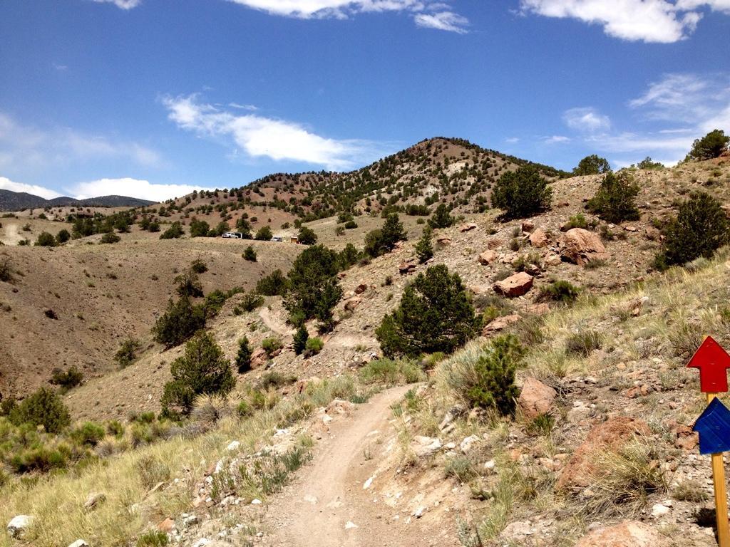A scenic view of a rugged landscape featuring rolling hills and sparse vegetation. The foreground shows a dirt path meandering through the terrain, with colorful trail markers indicating direction. In the background, distant mountains rise under a partly cloudy sky. The area is dotted with small trees and rocky outcrops, suggesting a natural and wild environment. Lil Rattler mountain bike trail.