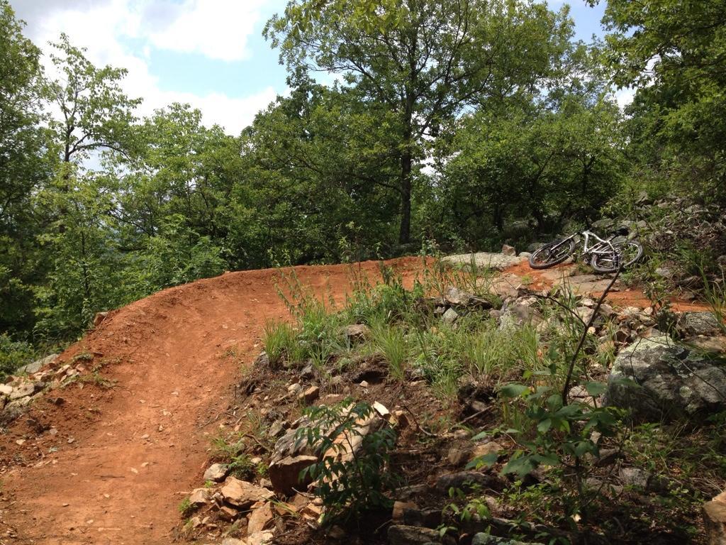A dirt mountain biking trail winding through a wooded area, with lush greenery on either side. A white mountain bike lies on the ground near the trail, partially obscured by rocks and grass. The sky above is partly cloudy. Coldwater Mountain mountain bike trail.
