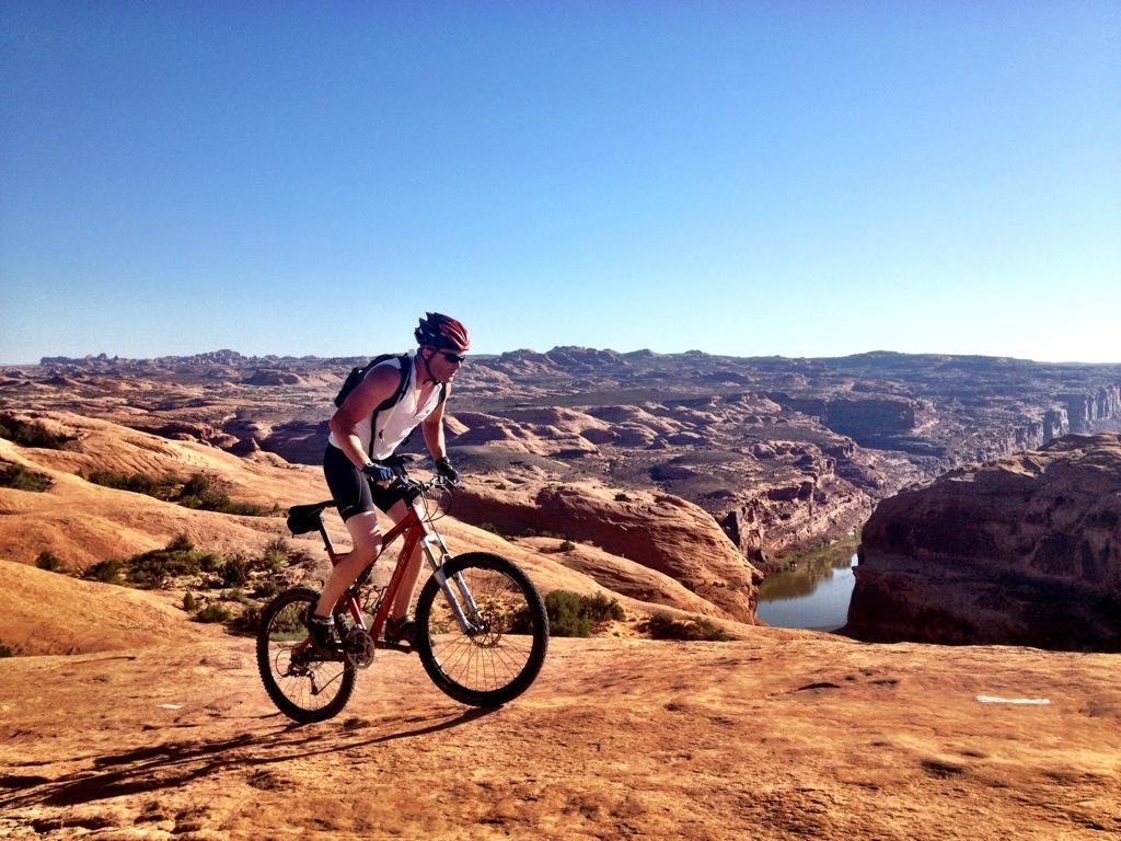 A mountain biker riding on rocky terrain with a scenic view of canyons and a river in the background under a clear blue sky. Slickrock mountain bike trail.