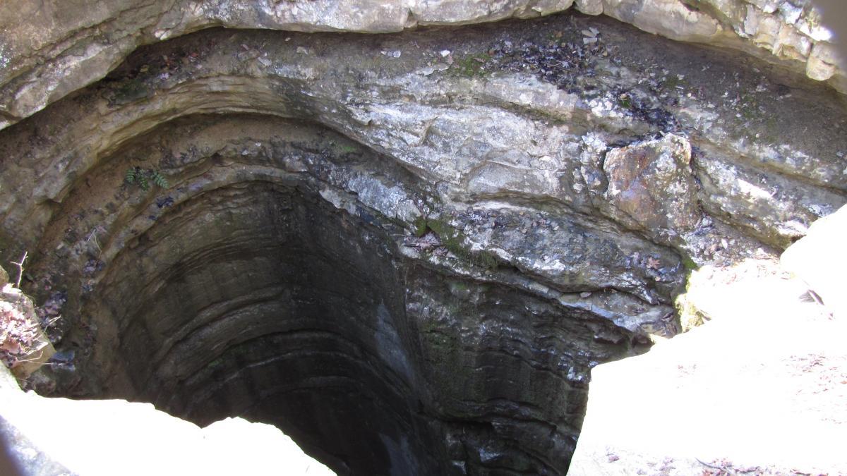 Aerial view of a deep, circular limestone sinkhole showing layers of rock and soil, surrounded by uneven terrain and sparse vegetation. Monte Sano State Park &amp; Land Trust mountain bike trail.