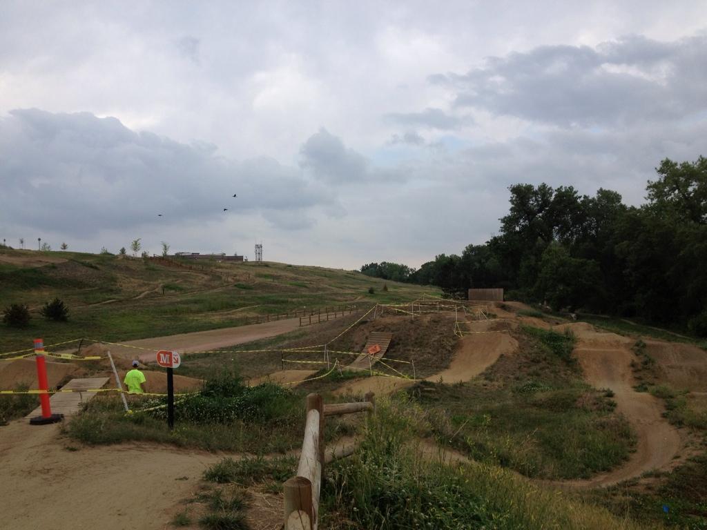 A scenic view of a dirt bike trail featuring ramps and jumps, surrounded by grassy hills and trees under a cloudy sky. A person in a bright green shirt stands near a sign, indicating a trail entrance, while yellow caution tape outlines the area. Valmont Bike Park mountain bike trail.