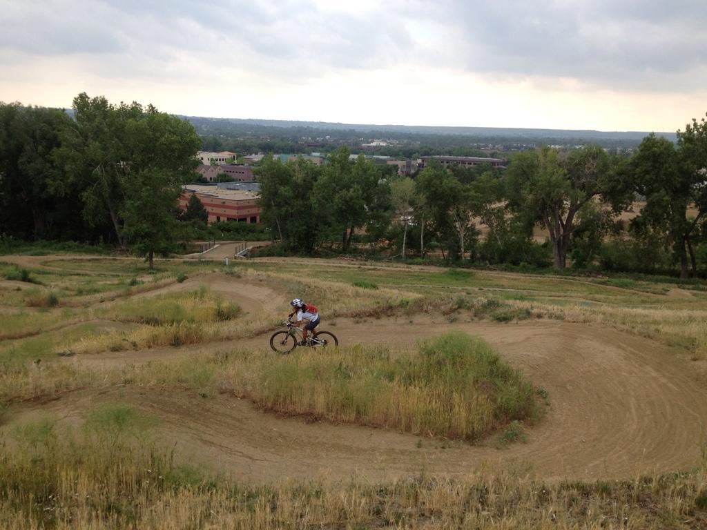 A cyclist rides on a dirt path with grassy embankments in a hilly area. In the background, a cityscape is visible with buildings and trees, under a cloudy sky. Valmont Bike Park mountain bike trail.