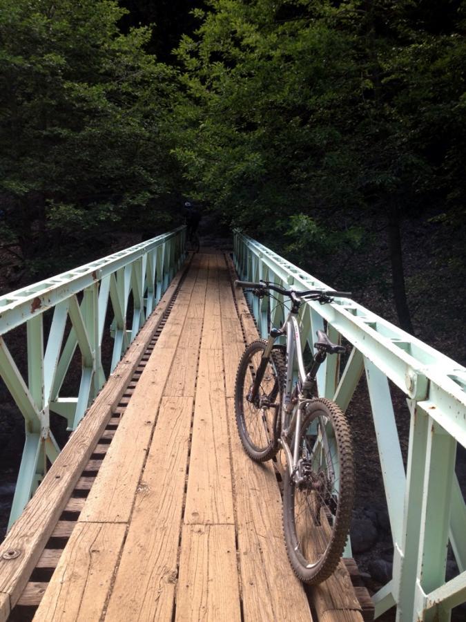 A mountain bike is parked on a wooden bridge surrounded by lush green trees. The bridge has a light blue metal frame and leads into a dense forest, creating an inviting path for outdoor adventures. Downieville Downhill mountain bike trail.