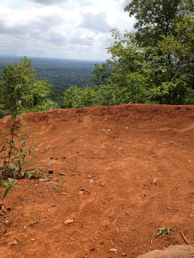 A view from a rocky cliff edge overlooking a lush, green valley, with a foreground of reddish-brown soil and sparse vegetation. The sky is partly cloudy, creating a scenic outdoor landscape. Coldwater Mountain mountain bike trail.