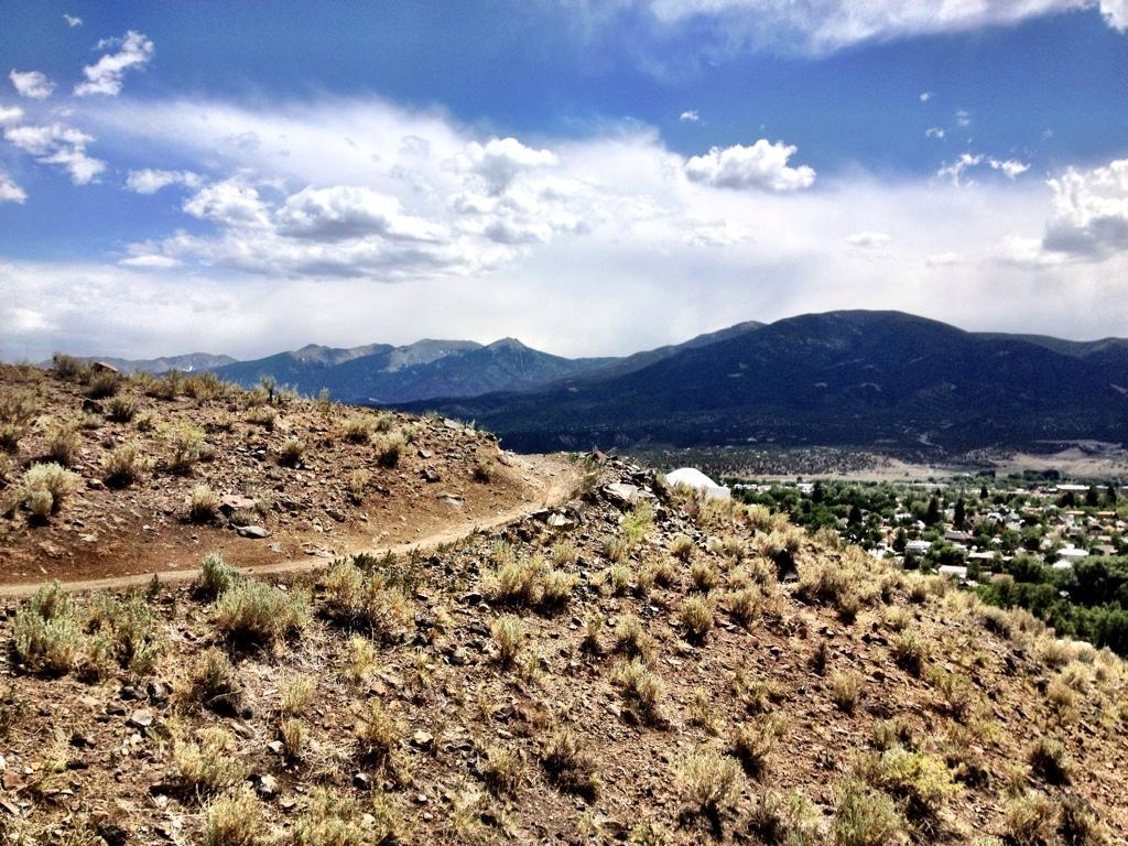 A scenic view from a rocky hillside overlooking a valley, with mountains in the background and a partly cloudy sky. The foreground features dry grass and small shrubs on the slope. The town is visible in the distance at the base of the mountains. Frontside mountain bike trail.