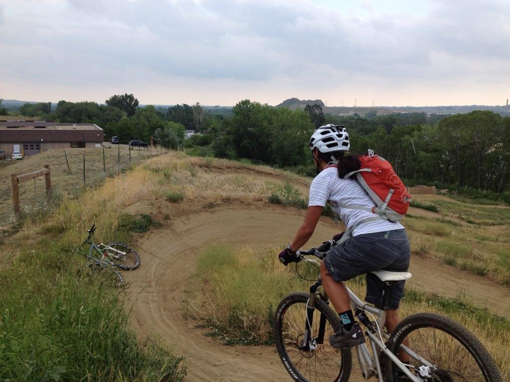 A person riding a mountain bike on a dirt trail, with a hilly landscape in the background. The biker is wearing a helmet and a backpack, and there is a bicycle leaning against the grass nearby. The sky is overcast, suggesting a cloudy day. Valmont Bike Park mountain bike trail.