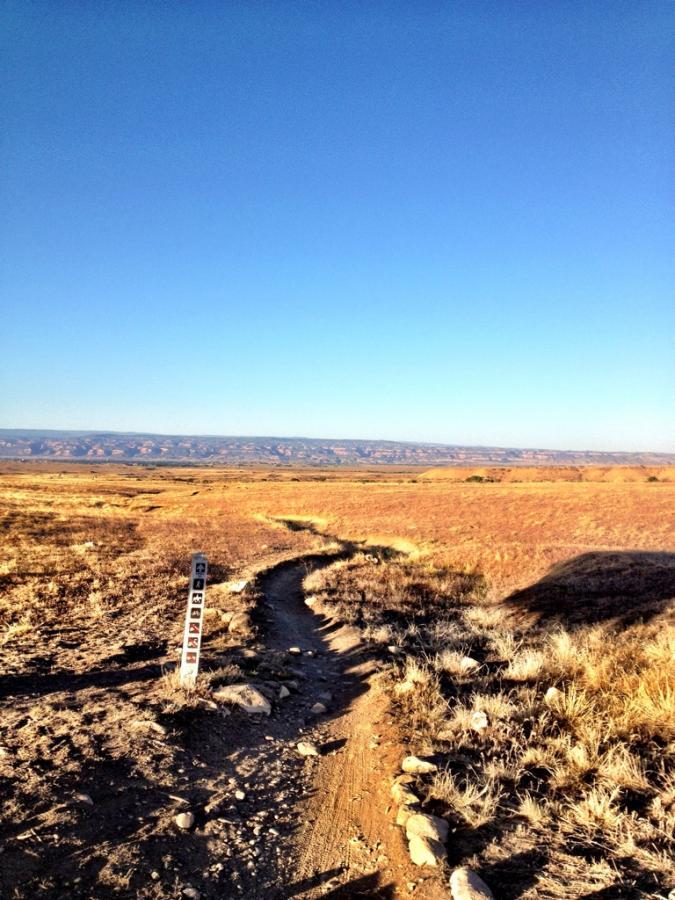 A winding dirt trail leads through a wide, open landscape of dry grasses and sparse vegetation, under a clear blue sky. In the foreground, a trail marker stands beside the path, indicating directions. The distant hills are visible on the horizon, complementing the serene and expansive natural setting. Kessel Run mountain bike trail.