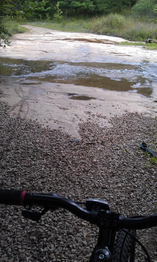 A view from the handlebars of a mountain bike, overlooking a rocky path that leads to a shallow stream. The ground is covered in small pebbles and patches of grass surround the area, with trees visible in the background. Harbins Park mountain bike trail.