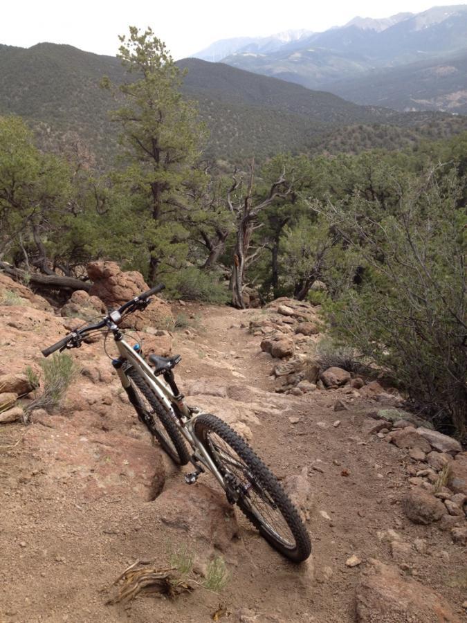 A mountain bike rests on a rocky trail leading down a mountainside, surrounded by trees and shrubs. In the background, rolling hills and distant mountains are visible under a cloudy sky. Unkle Nazty mountain bike trail.