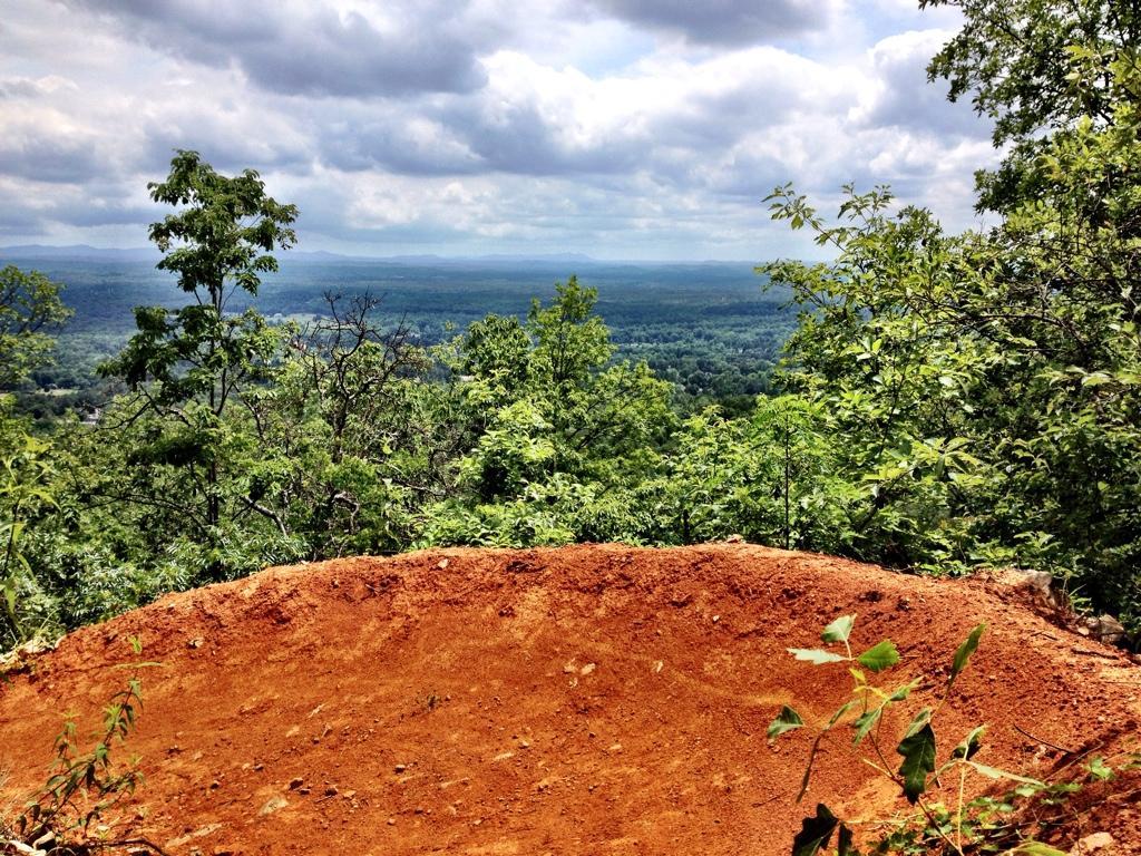 A scenic view from a rocky ledge overlooking a lush green landscape with rolling hills under a cloudy sky. The foreground features a reddish-brown earth patch, surrounded by various trees and foliage. Coldwater Mountain mountain bike trail.
