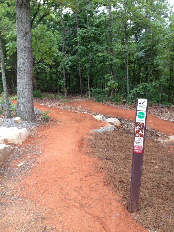 A winding dirt trail through a wooded area, marked by a sign indicating trail usage. The path is surrounded by trees and features a reddish-brown dirt surface, with some rocks and pine needles along the edges. Coldwater Mountain mountain bike trail.
