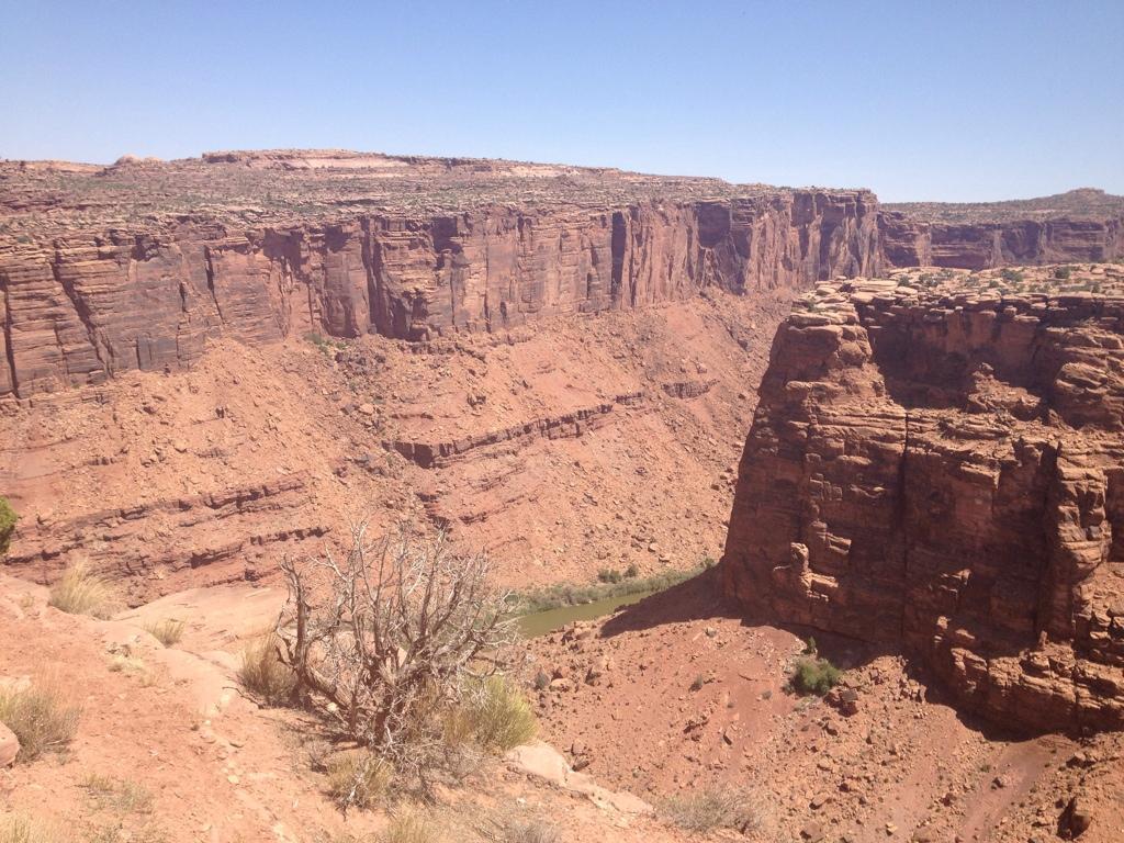 A wide view of a rugged canyon landscape featuring steep reddish cliffs, with a narrow river winding through the valley below. The sky is clear and bright, showcasing a natural, arid environment with sparse vegetation. Porcupine Rim mountain bike trail.