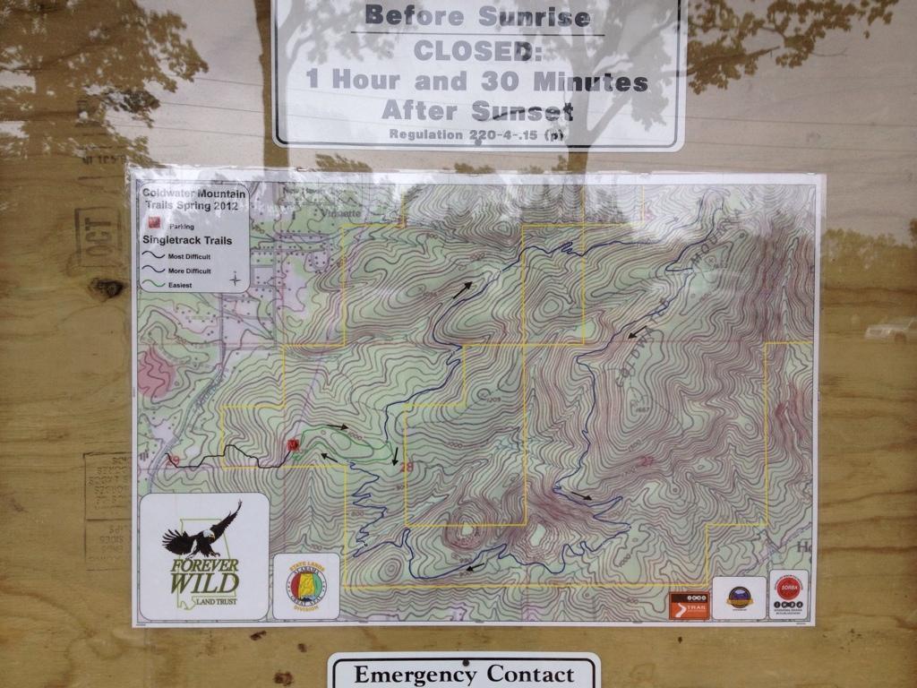 Map of Coldwater Mountain trails, featuring various singletrack trails labeled by difficulty level (most difficult, more difficult, easiest). The map includes a parking area icon and emergency contact information, with a visible notice stating the hours of trail closure before sunrise and after sunset. Coldwater Mountain mountain bike trail.
