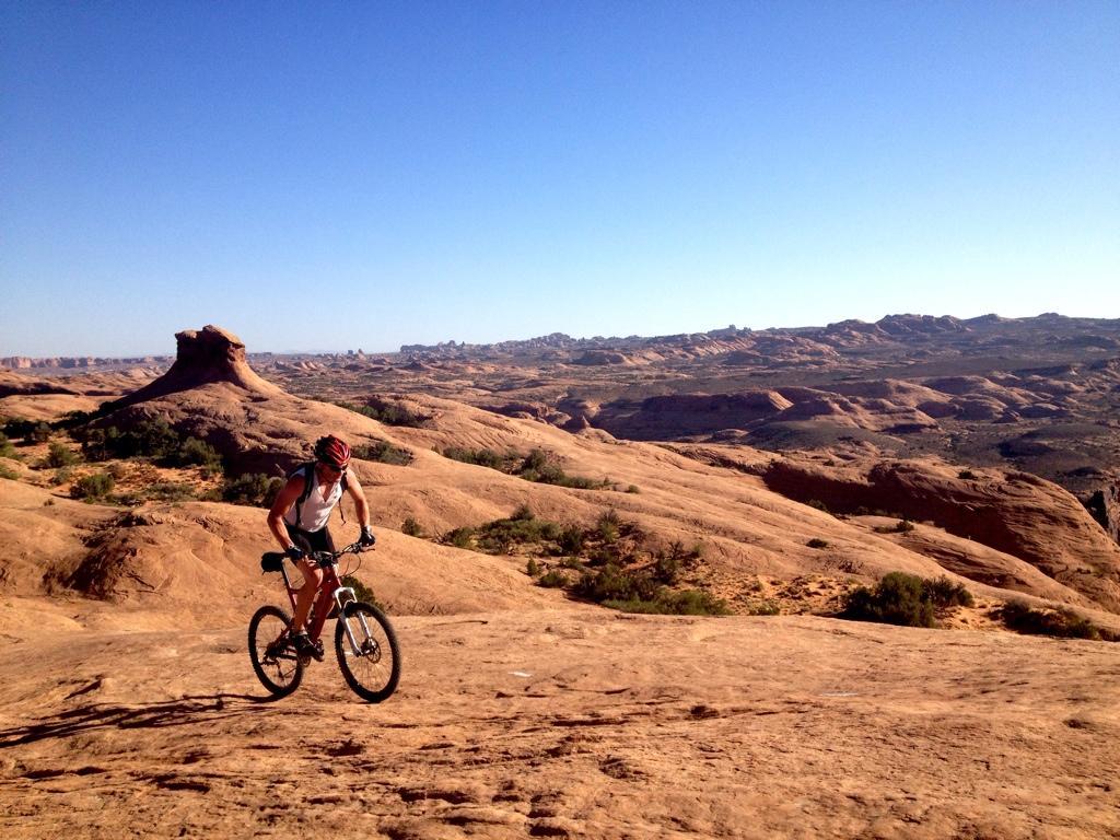 A mountain biker navigates a rocky terrain with a clear blue sky overhead, surrounded by expansive desert landscapes and unique rock formations. Slickrock mountain bike trail.