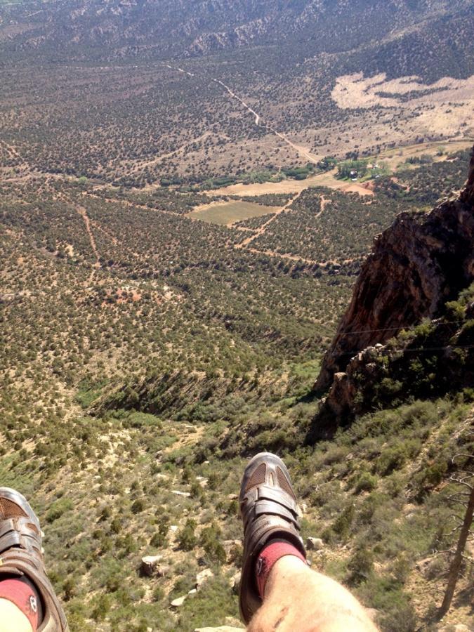 Feet in climbing shoes dangle over a rocky cliff edge, overlooking a vast landscape of green and brown terrain. The view captures rolling hills, patches of vegetation, and clear skies in the distance. Porcupine Rim mountain bike trail.