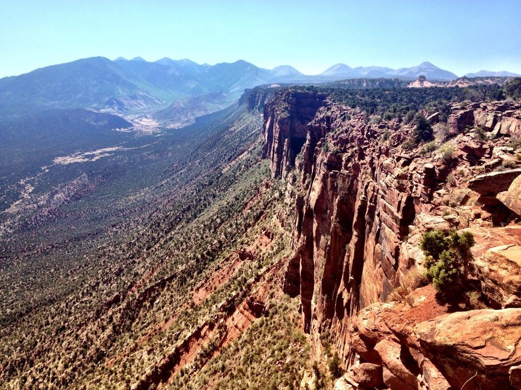 A panoramic view of a rugged cliffside landscape featuring red rock formations and lush greenery, with distant mountains visible under a clear blue sky. The steep cliffs drop dramatically into a valley below, showcasing the natural beauty of the terrain. Porcupine Rim mountain bike trail.