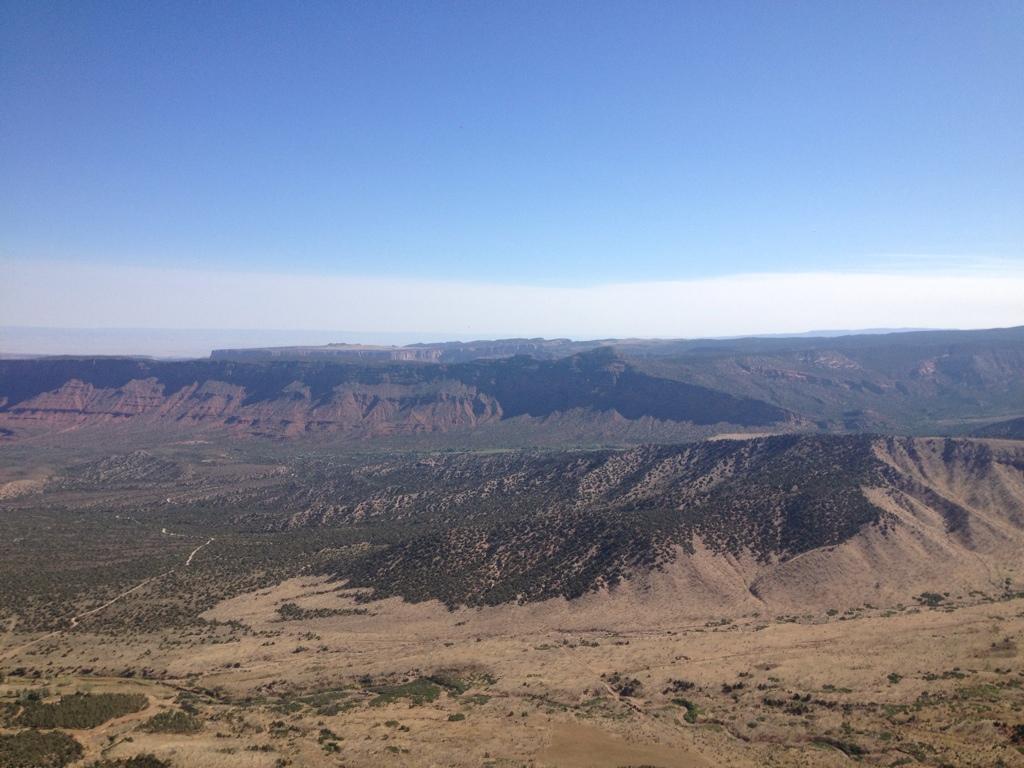 A panoramic view of a rugged mountainous landscape under a clear blue sky, showcasing rolling hills, steep cliffs, and a mix of vegetation and bare earth. The distant mountains provide a dramatic backdrop to the serene foreground of scrubland and winding paths. UPS And LPS mountain bike trail.