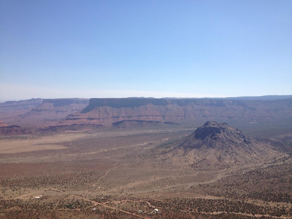 A panoramic view of a desert landscape featuring rugged cliffs and mesas under a clear blue sky. In the foreground, a prominent rock formation rises sharply from the flat terrain, surrounded by sparse vegetation. Porcupine Rim mountain bike trail.