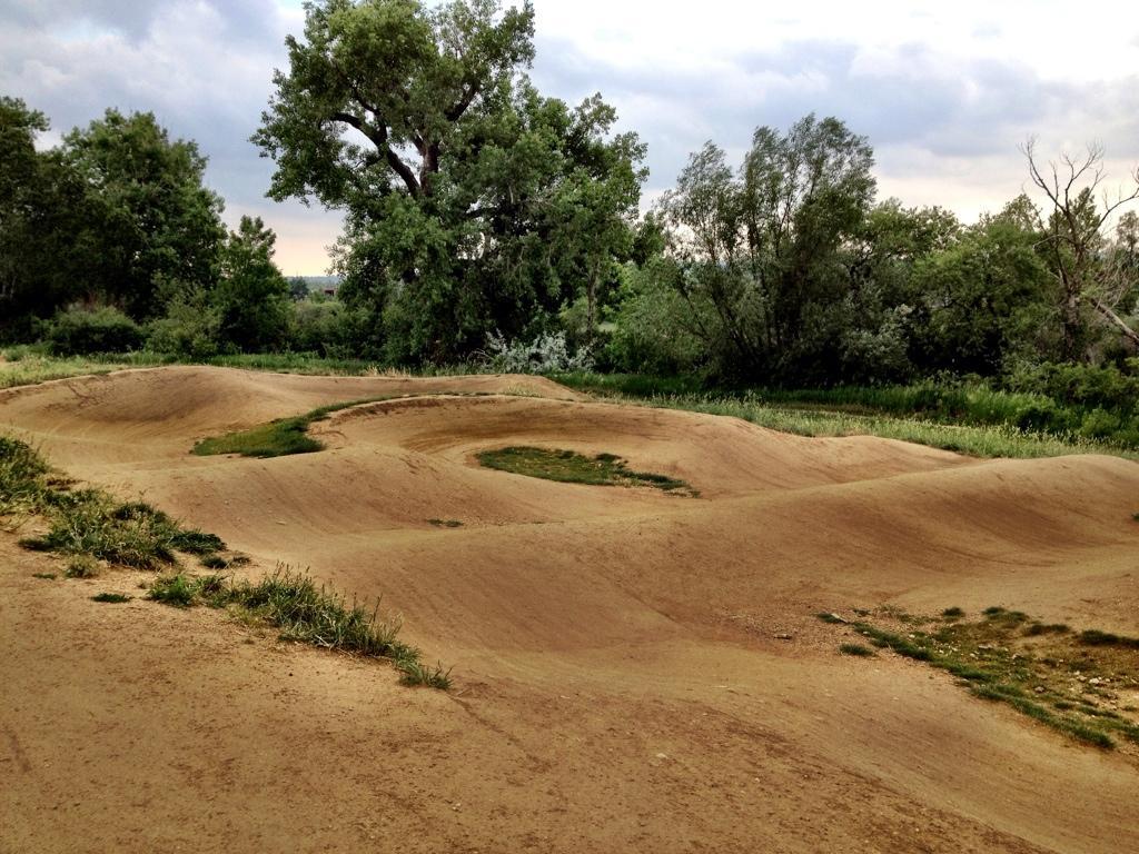 A dirt bike pump track with undulating mounds and curves, surrounded by lush greenery and trees under a partly cloudy sky. Valmont Bike Park mountain bike trail.