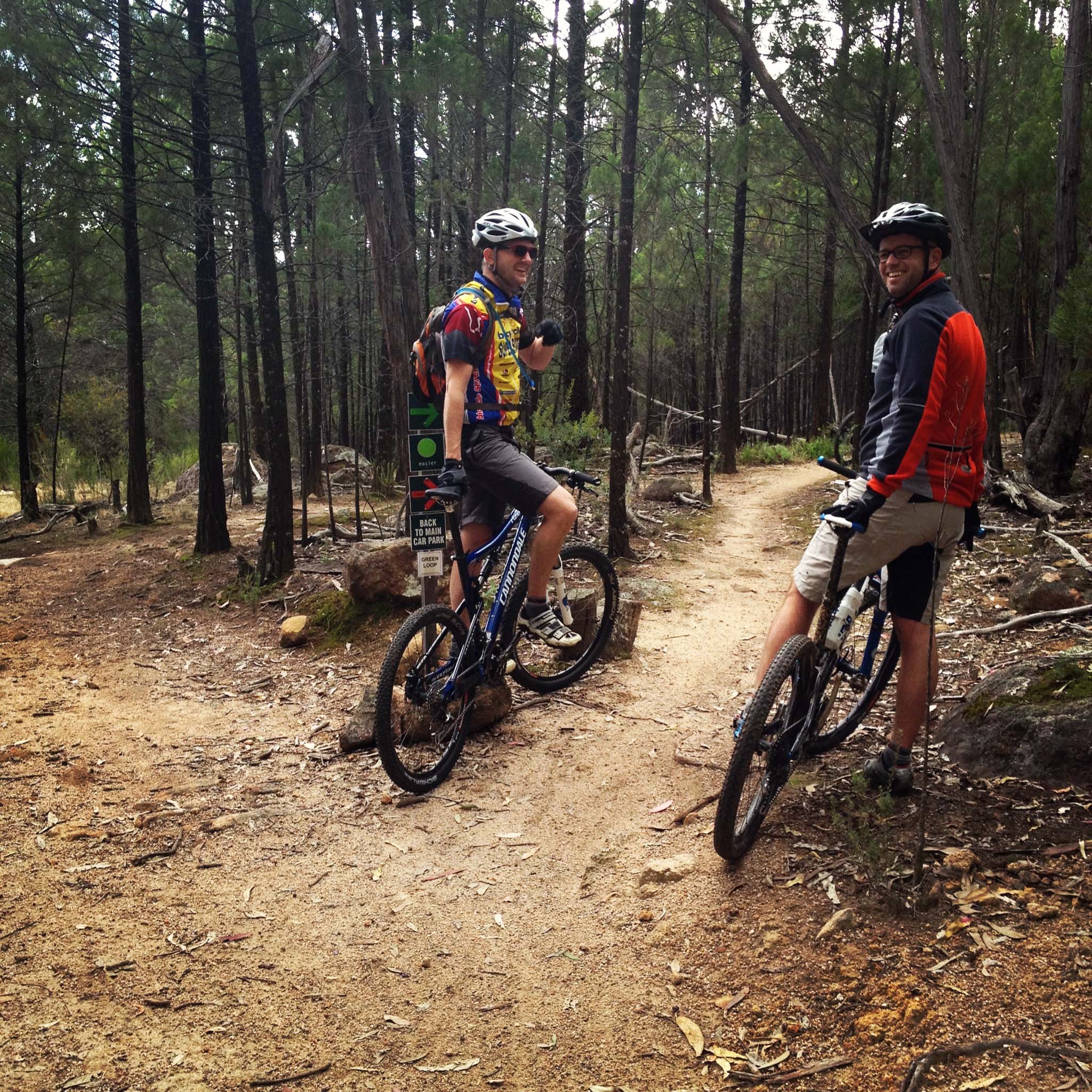 Two mountain bikers on a dirt trail in a wooded area, wearing helmets and biking gear. One rider is giving a thumbs up while the other is looking back, both positioned near a fork in the trail surrounded by trees.