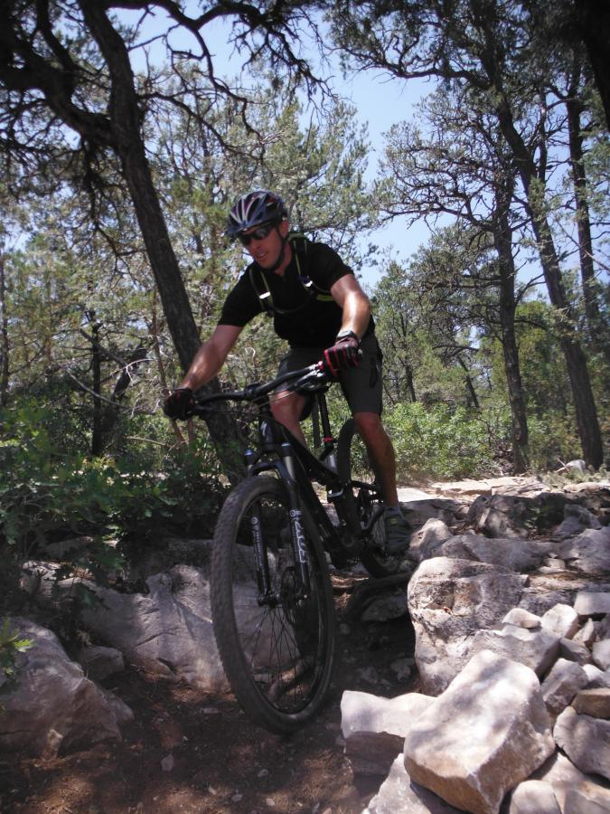 Mountain biker navigating rocky terrain in a forested area, wearing a helmet and cycling gear. Sunlight filters through the trees in the background. Otero Canyon mountain bike trail.