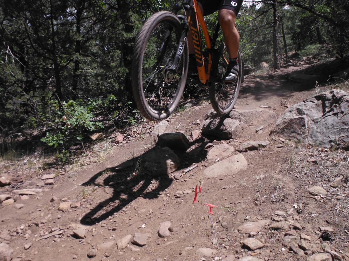 A mountain biker jumps over a rock on a dirt trail surrounded by trees, with dust kicking up from the ground. Bright red markers are visible on the trail in the foreground. Otero Canyon mountain bike trail.