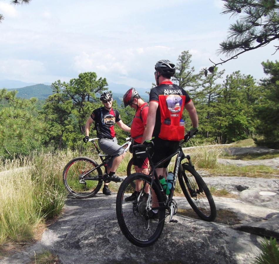 Three mountain bikers standing on rocky terrain, wearing helmets and cycling gear, while discussing their ride in a scenic outdoor setting with trees and mountains in the background. Cedar Rock Trail #16 mountain bike trail.