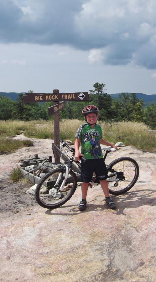 A young boy wearing a helmet and a colorful shirt stands beside his mountain bike at a trail sign labeled "Big Rock Trail." The backdrop features a scenic view of grassy hills and distant mountains under a partly cloudy sky. Cedar Rock Trail #16 mountain bike trail.