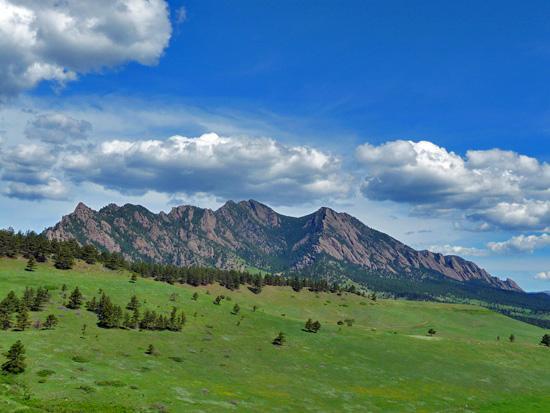 A scenic view of a mountain range under a blue sky with scattered clouds, surrounded by a green landscape with patches of grass and small trees in the foreground. Doudy Draw Trail mountain bike trail.