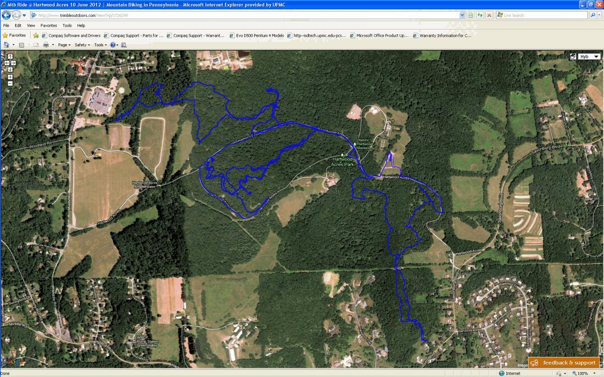 A satellite image map showing a trail route marked in blue, located within Hartwood Acres Park in Pennsylvania. The map displays various curves and paths winding through wooded areas, fields, and nearby roads, providing an overview suitable for mountain biking or outdoor activities. Hartwood Acres mountain bike trail.