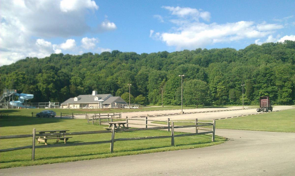 A scenic view of a park area featuring a building, picnic tables, and a water slide in the foreground, with a lush green hillside in the background under a partly cloudy sky. A parked car and a truck can be seen in the empty parking lot. Versailles State Park mountain bike trail.