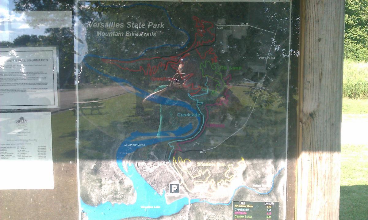 Map of Versailles State Park displaying mountain bike trails, including Shadow Run, Creekside, and Center Loop. The map features trail lengths and other important park information, with a clear layout of the surrounding area and trails. A picnic area is visible in the background. Versailles State Park mountain bike trail.