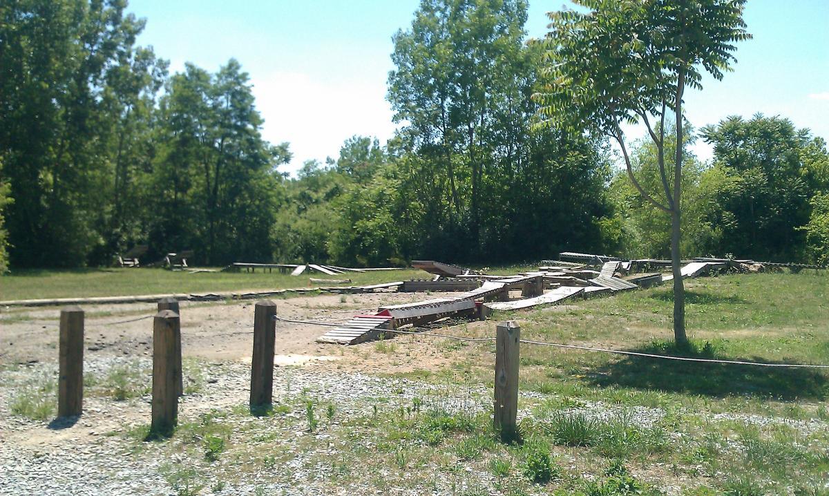 A view of a grassy area with a series of wooden ramps and structures, partially overgrown, set against a backdrop of trees under a clear blue sky. Wooden posts line the foreground, suggesting the boundary of the space. Rangeline Nature Preserve mountain bike trail.