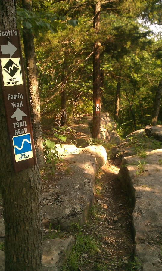 A wooded trail area featuring directional signs indicating "Scott's" trail, labeled as "Most Difficult," and a "Family Trail," marked as "More Difficult." The path is lined with rocks and greenery, leading into the forest. Landahl Park Reserve mountain bike trail.