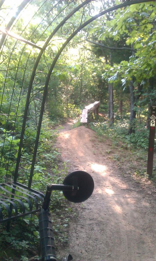 A view of a dirt biking trail entrance framed by a decorative archway, surrounded by lush greenery. In the background, a dirt jump ramp is visible along the trail, with a trail marker sign nearby. The scene is illuminated by soft sunlight filtering through the trees. Landahl Park Reserve mountain bike trail.