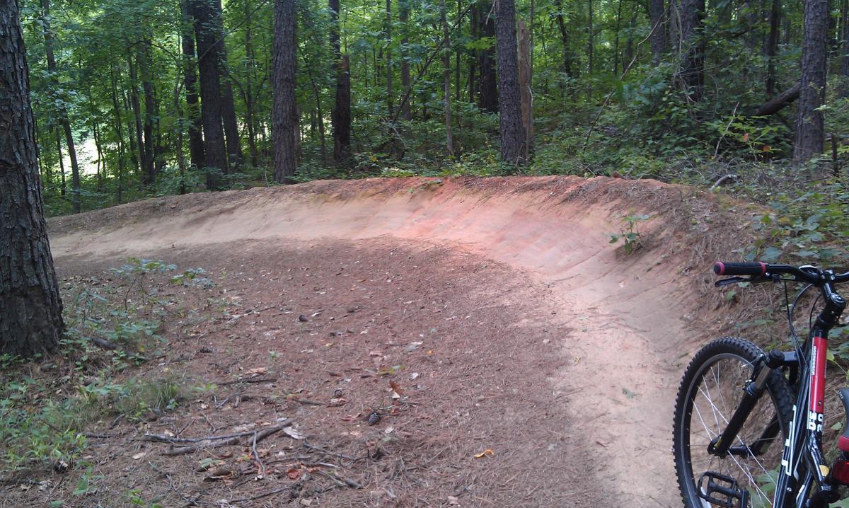 A winding dirt bike trail in a wooded area, with a mountain bike resting on the right side. The trail features a curved path with reddish soil, surrounded by tall trees and lush greenery. Harbins Park mountain bike trail.