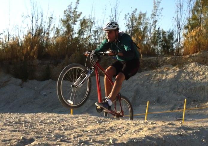 A male cyclist performing a jump on a mountain bike over a dirt track, with a backdrop of trees and a clear sky. The cyclist is wearing a helmet and sports attire, demonstrating an active outdoor sport. Virginia Key North Point mountain bike trail.