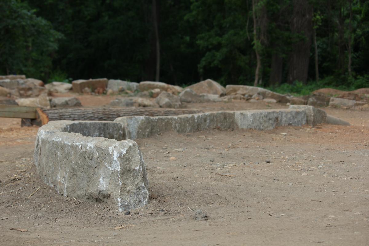 A landscape featuring a section of large, weathered stones arranged in a curved formation on a dirt surface. Surrounding the stones are various other rocks and sparse vegetation, indicating a natural setting. The background includes dense green trees, contributing to an overall impression of an archaeological site or historical location. Belle Isle mountain bike trail.
